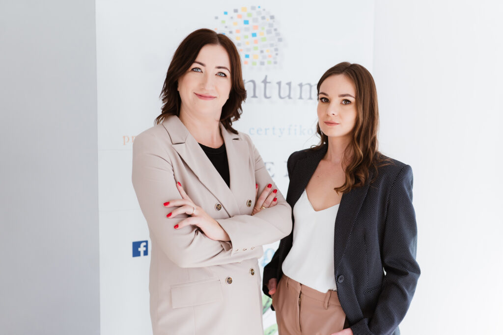 Dwie kobiety w profesjonalnych strojach stoją obok siebie, uśmiechając się pewnie. Jedna z nich ma skrzyżowane ręce i nosi beżową marynarkę, podczas gdy druga ma na sobie ciemną marynarkę i trzyma rękę w kieszeni. W tle znajduje się niewyraźne logo firmy.