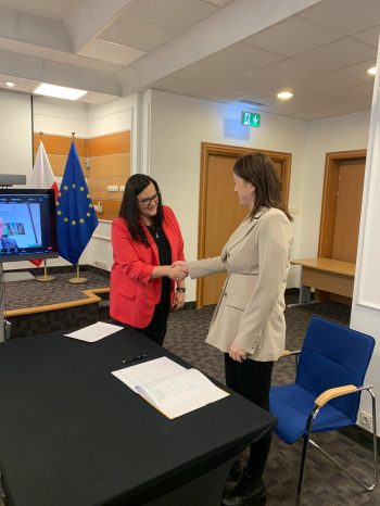 Dwie kobiety ściskają sobie dłonie w otoczeniu biurowym. Na pierwszym planie znajduje się st&oacute;ł z dokumentami, a w tle ekran, flagi Polski i UE oraz krzesła. Obie kobiety są uśmiechnięte i ubrane w stroje biznesowe.