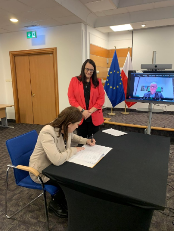 Kobieta siedzi przy stole i podpisuje dokument, podczas gdy inna kobieta stoi obok niej. Za nimi ekran pokazuje uczestnika połączenia wideo. W tle widoczne są flagi UE i flagi narodowe.